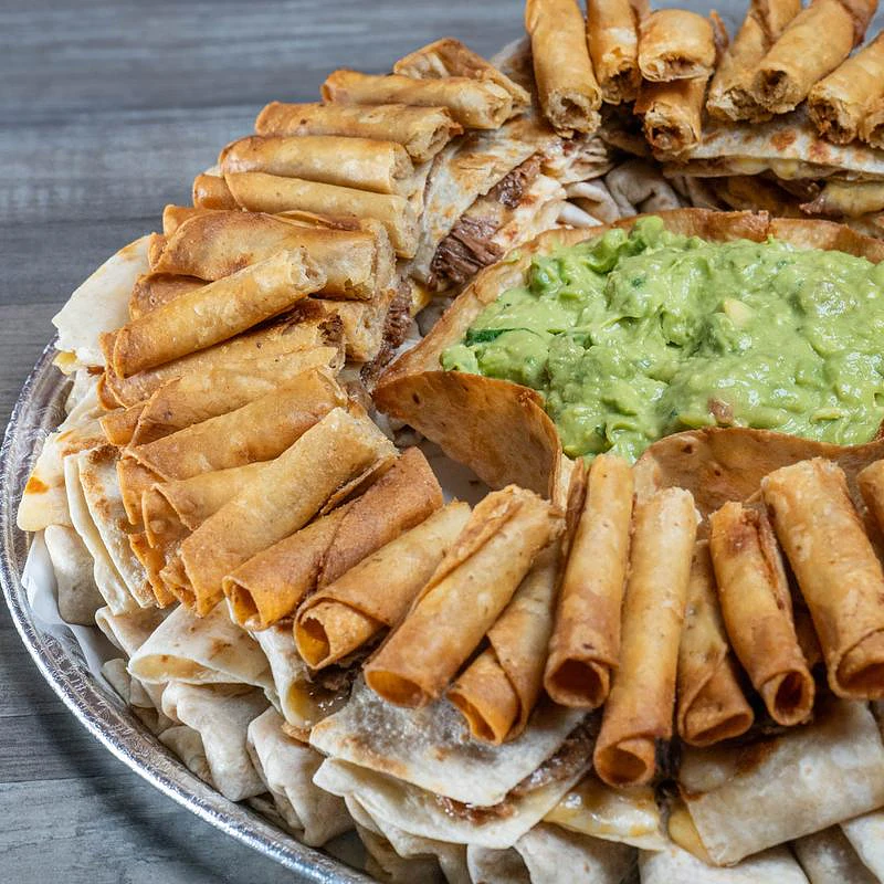Party tray of mini taquitos, burritos and quesadillas around a bowl of guacamole
